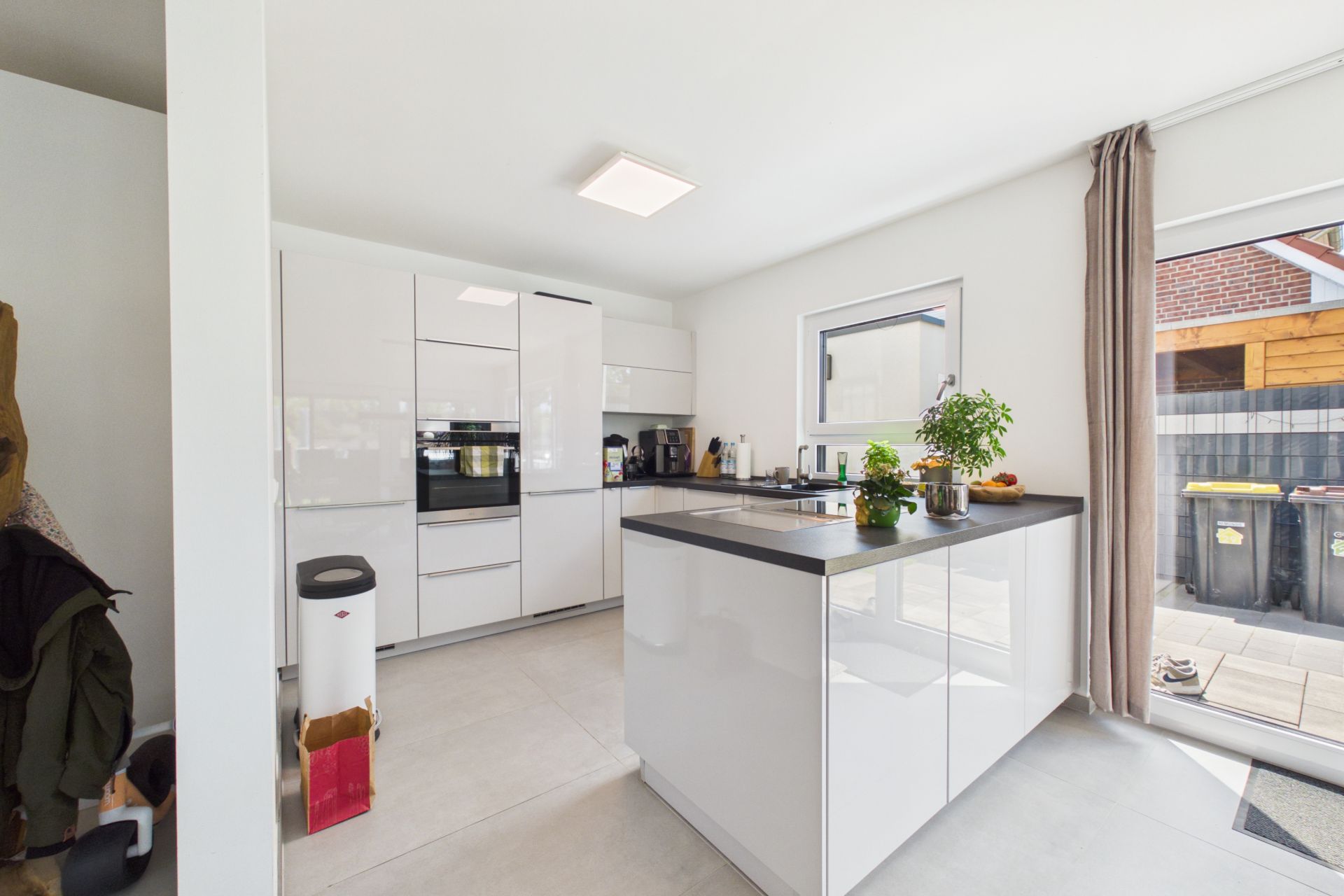 Moderne weiße Küche mit Kochinsel, Fenster und Zugang zur Terrasse, Pflanzen auf der Arbeitsfläche.