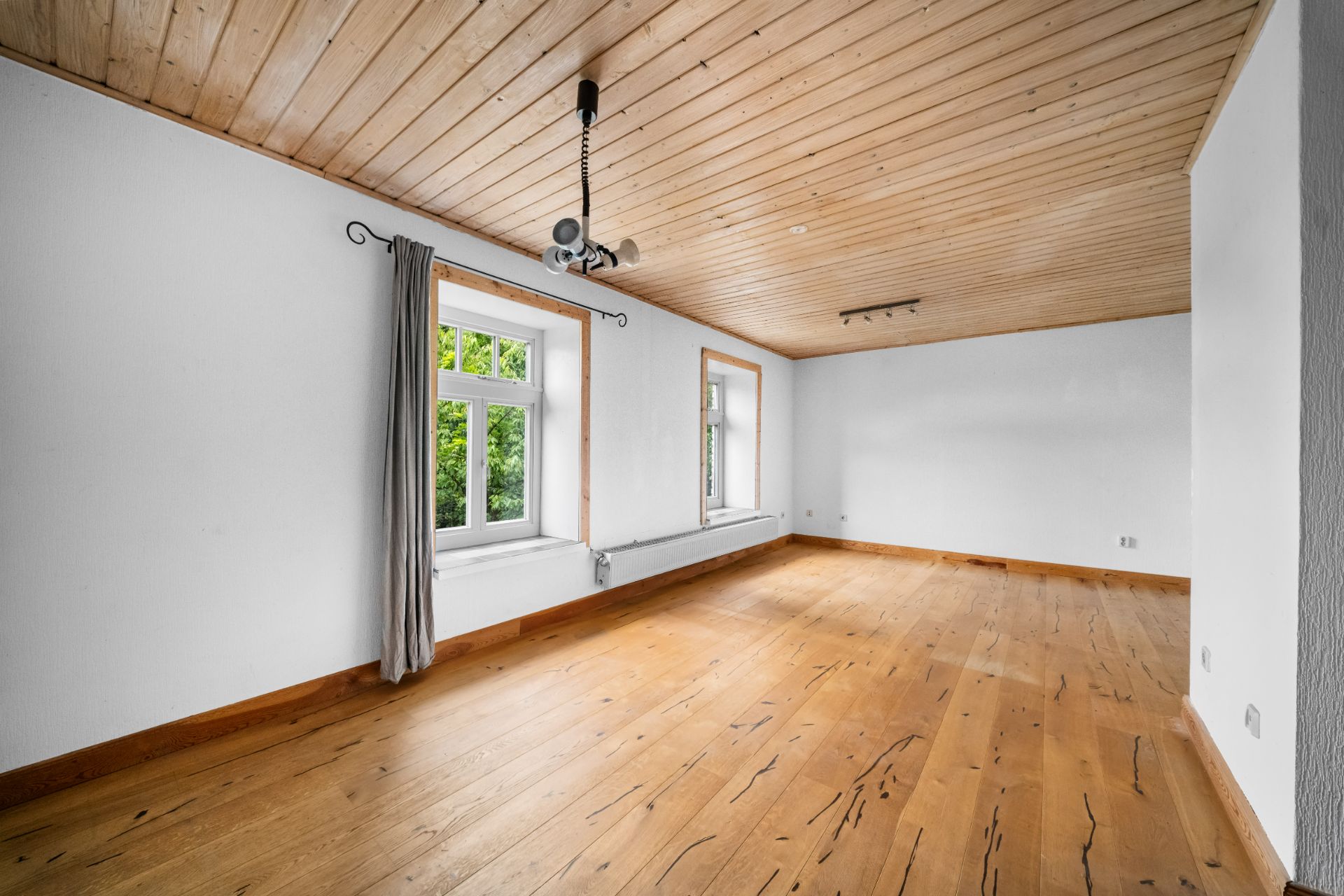 Leerer Raum mit Holzboden, Holzdecke, zwei Fenstern und einem Deckenprojektor.