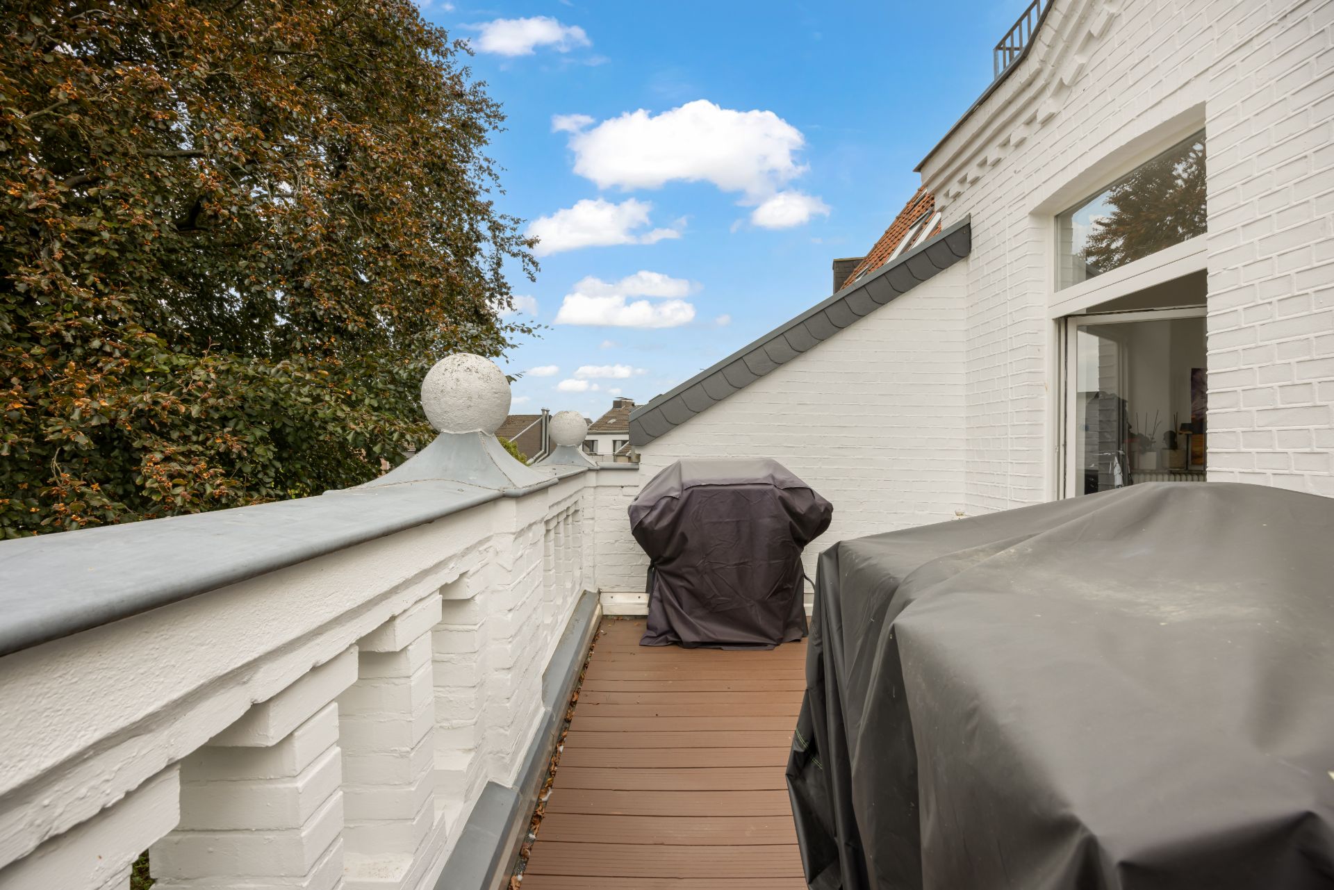 Balkon mit weißer Brüstung, zwei abgedeckte Gegenstände und Zugang zu einem Wohnbereich.