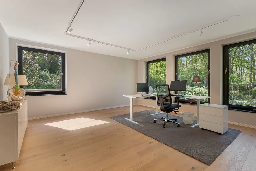 Helles Arbeitszimmer mit großem Schreibtisch, Stuhl und drei Fenstern mit Blick ins Grüne.