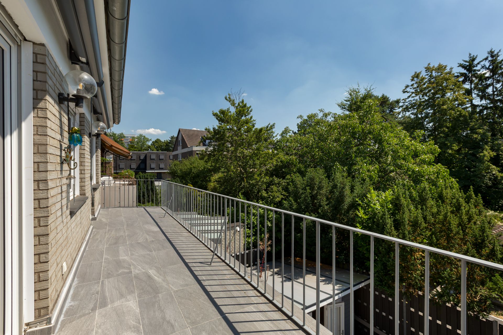 Großer Balkon mit grauem Boden und Metallgeländer, Blick auf Bäume und benachbarte Häuser.