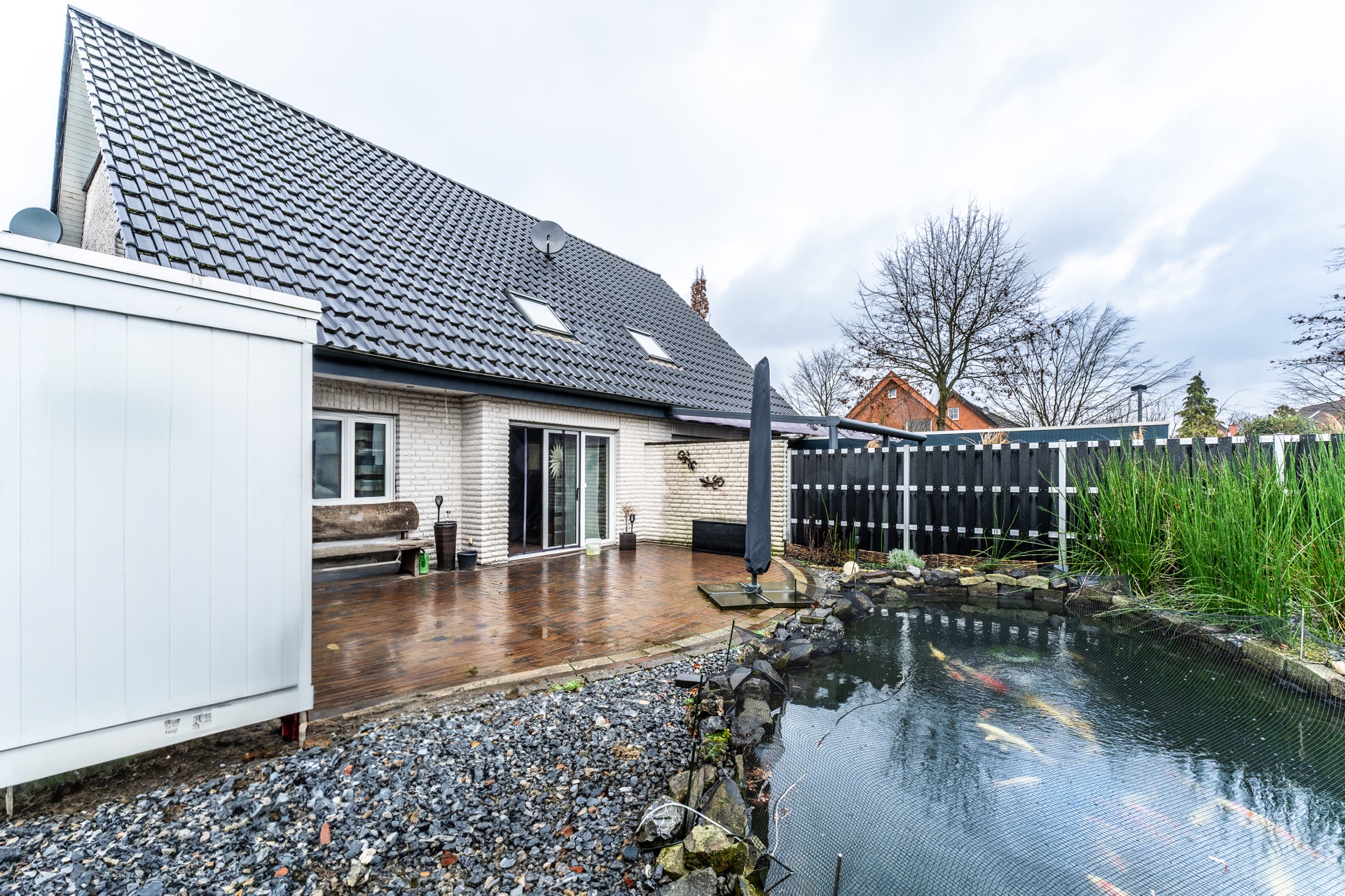 Gepflegter Garten mit Holzterrasse, Gartenbank und kleinem Teich neben einem Wohnhaus.