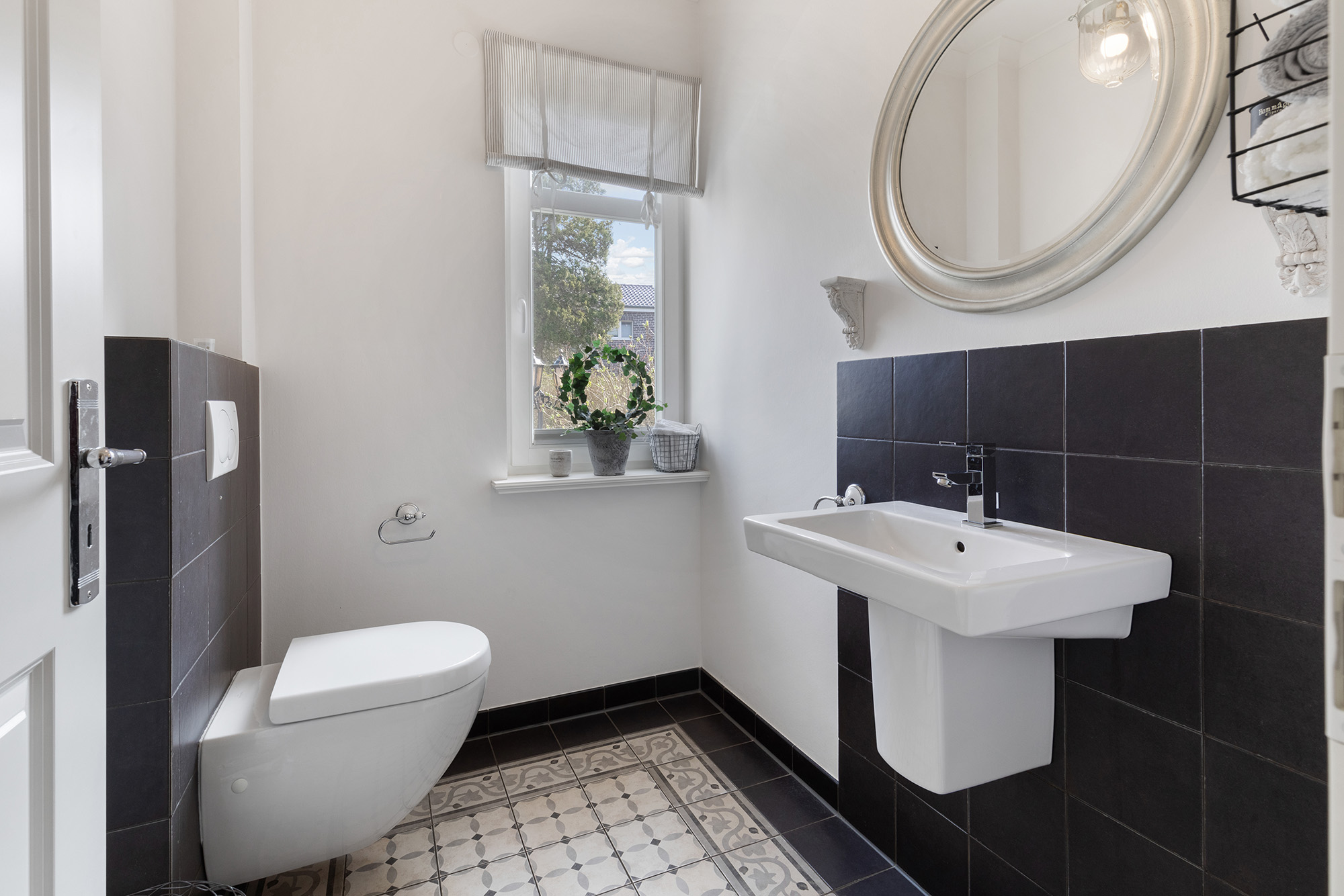 Helles Badezimmer mit Wandtoilette, Waschbecken, Spiegel und Fenster mit Pflanze auf der Fensterbank.