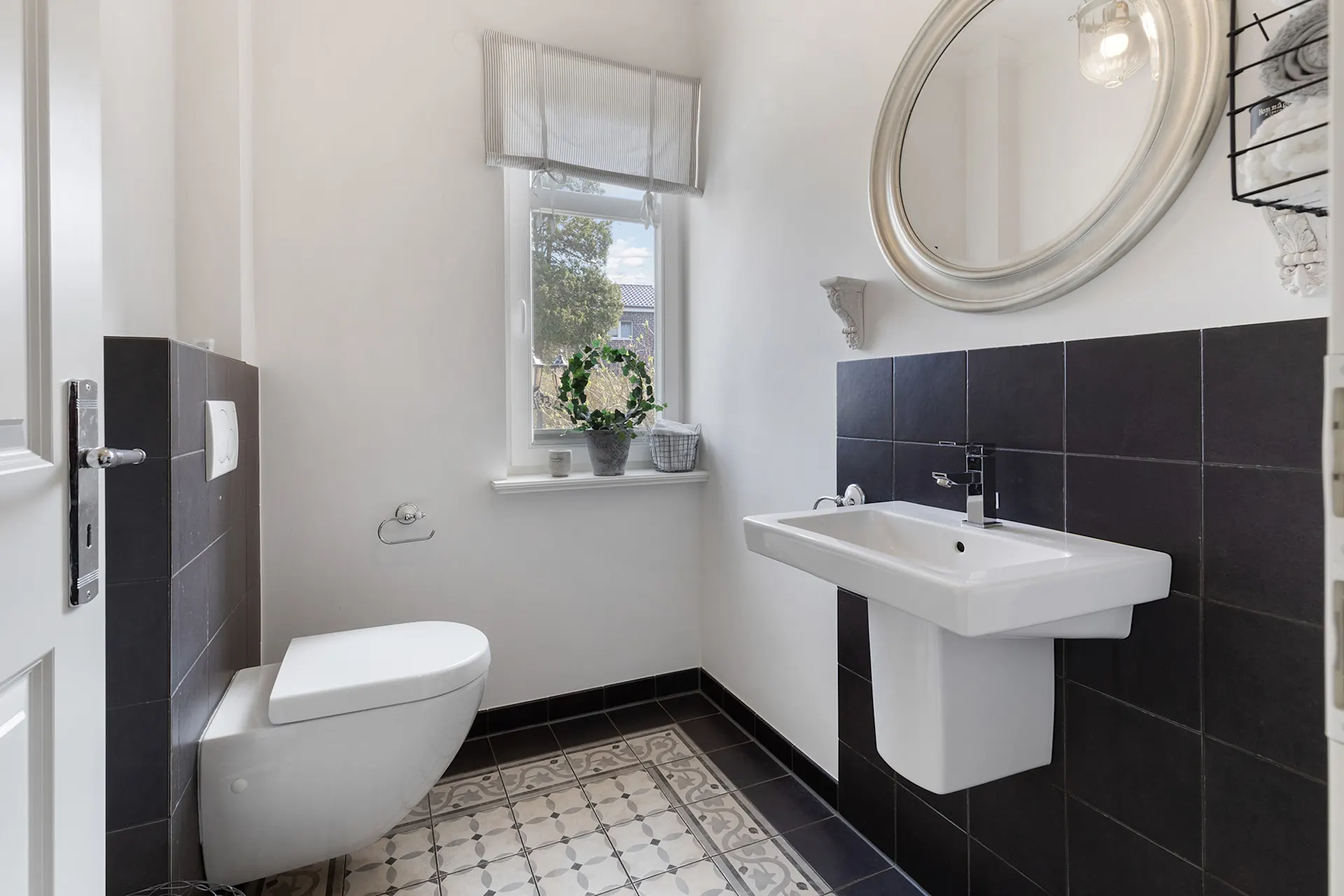 Helles Badezimmer mit Wandtoilette, Waschbecken, Spiegel und Fenster mit Pflanze auf der Fensterbank.