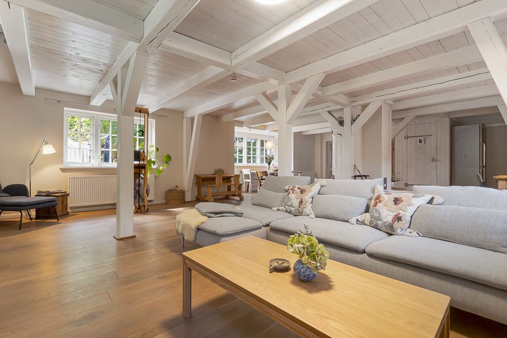 Helles Wohnzimmer mit großer grauer Couch, Holztisch und sichtbaren weißen Balken.