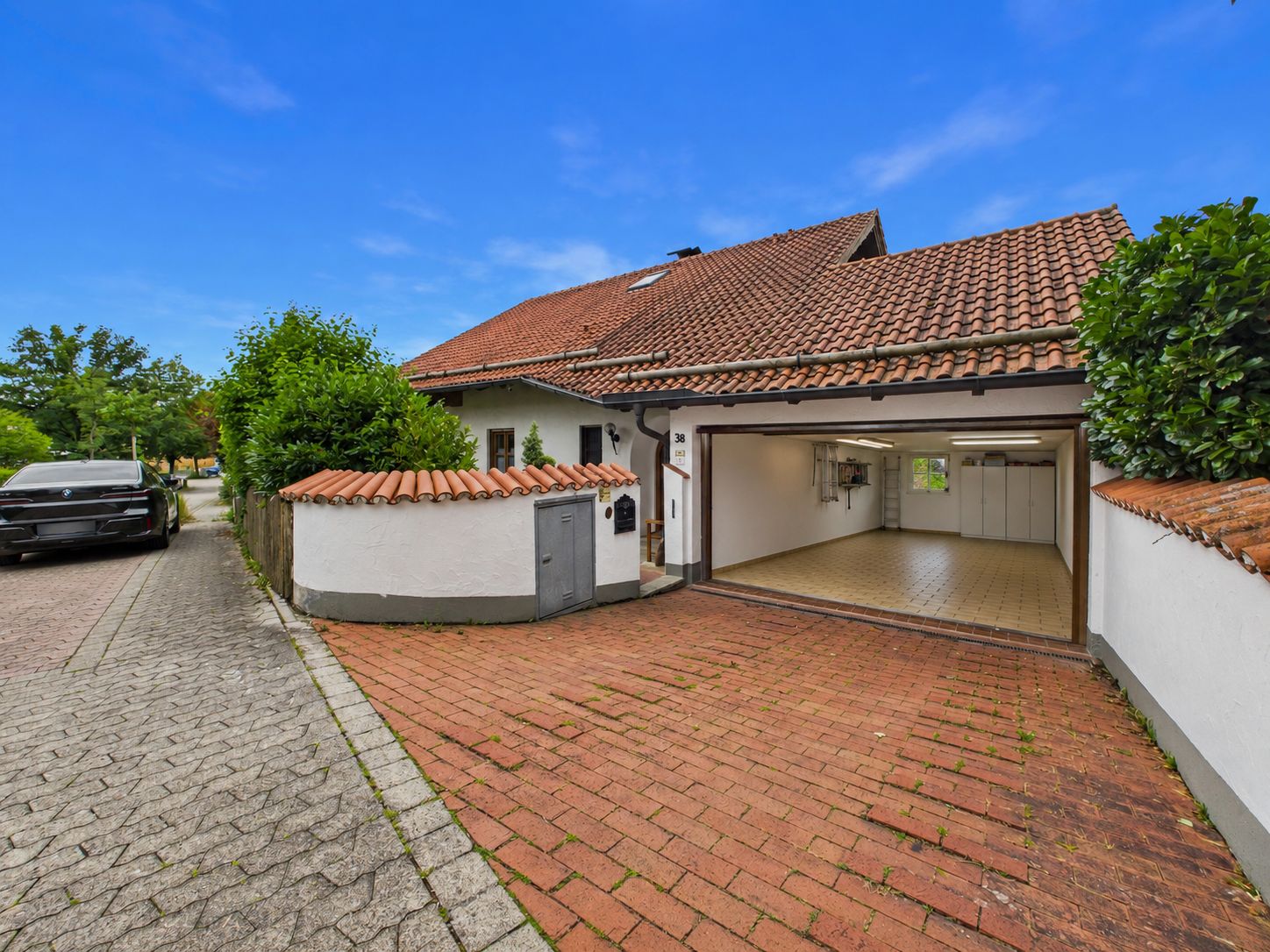 Offene Garage mit rotem Ziegeldach neben gepflasterter Auffahrt und schwarzem Auto bei Wohnhaus.