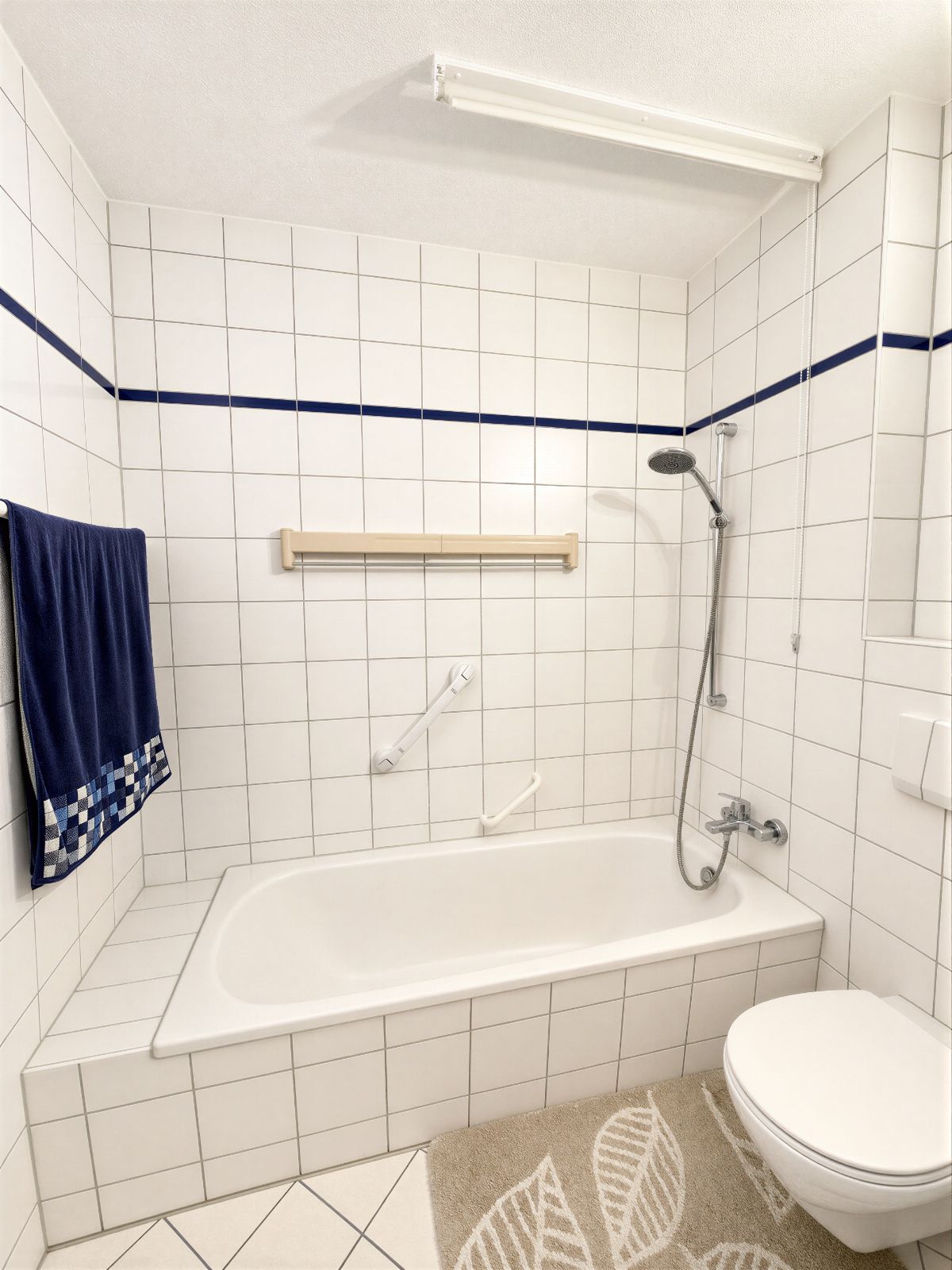 Weiß gefliestes Badezimmer mit Badewanne, Dusche, Haltegriffen, blauem Handtuch und WC.
