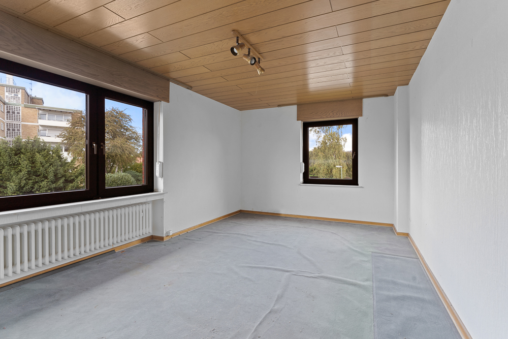 Leerer Raum mit hellem Holz an Decke, zwei Fenstern und weißem Heizkörper unter dem großen Fenster.