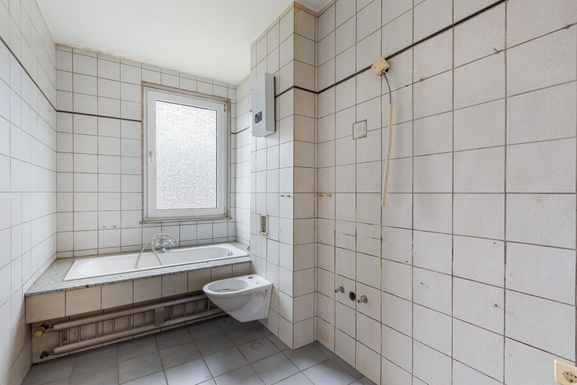 Leeres Badezimmer mit Badewanne, WC und Fenster, gefliest in Weiß.