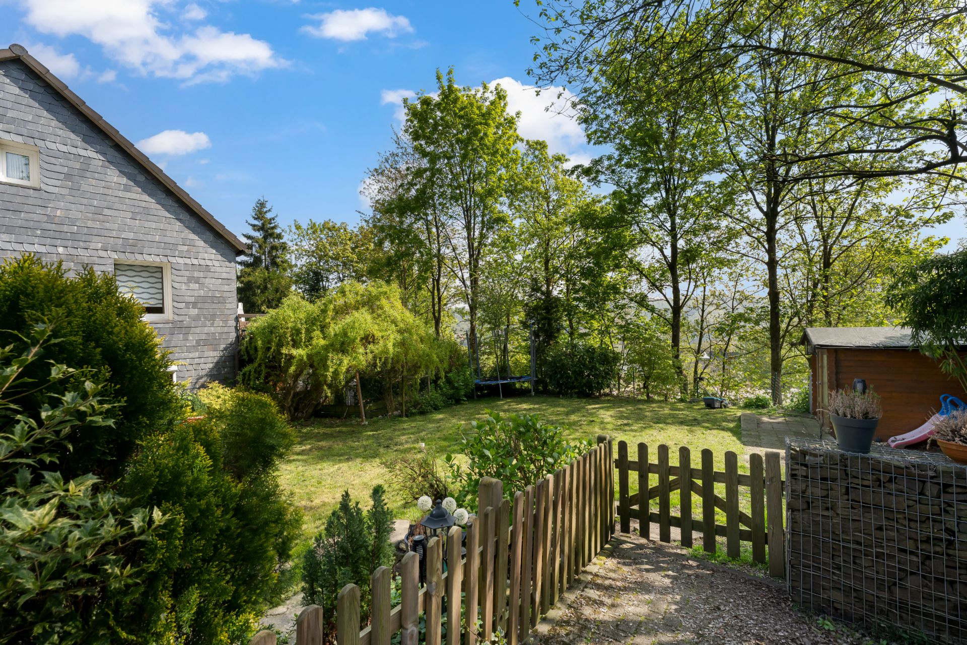 Garten mit Rasen, Holzzaun, Bäumen und kleinem Schuppen neben einem Wohnhaus.