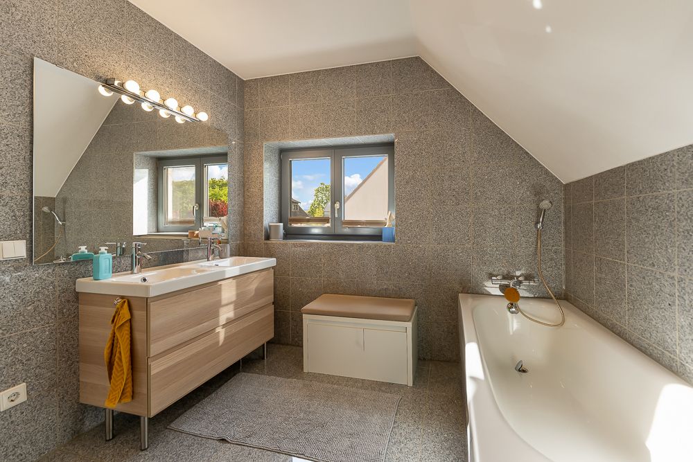 Modernes Badezimmer mit Doppelwaschbecken, Badewanne, Fenster und grauen Fliesen.