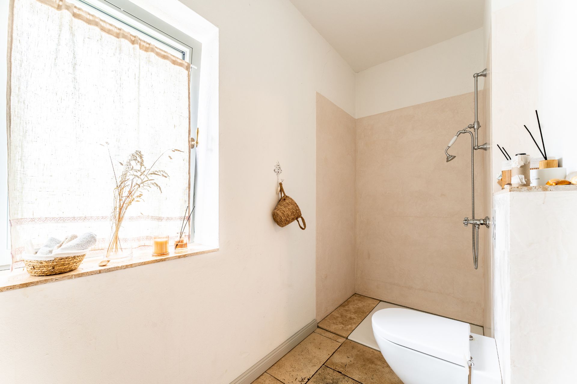 Helles Badezimmer mit ebenerdiger Dusche, Toilette, Fenster mit Vorhang und dekorativen Elementen.