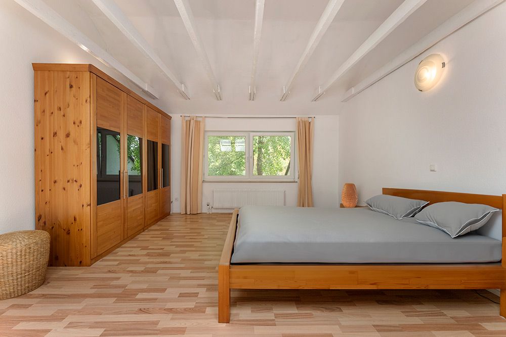 Helles Schlafzimmer mit Holzkleiderschrank, Doppelbett und Fenster mit Vorhängen.