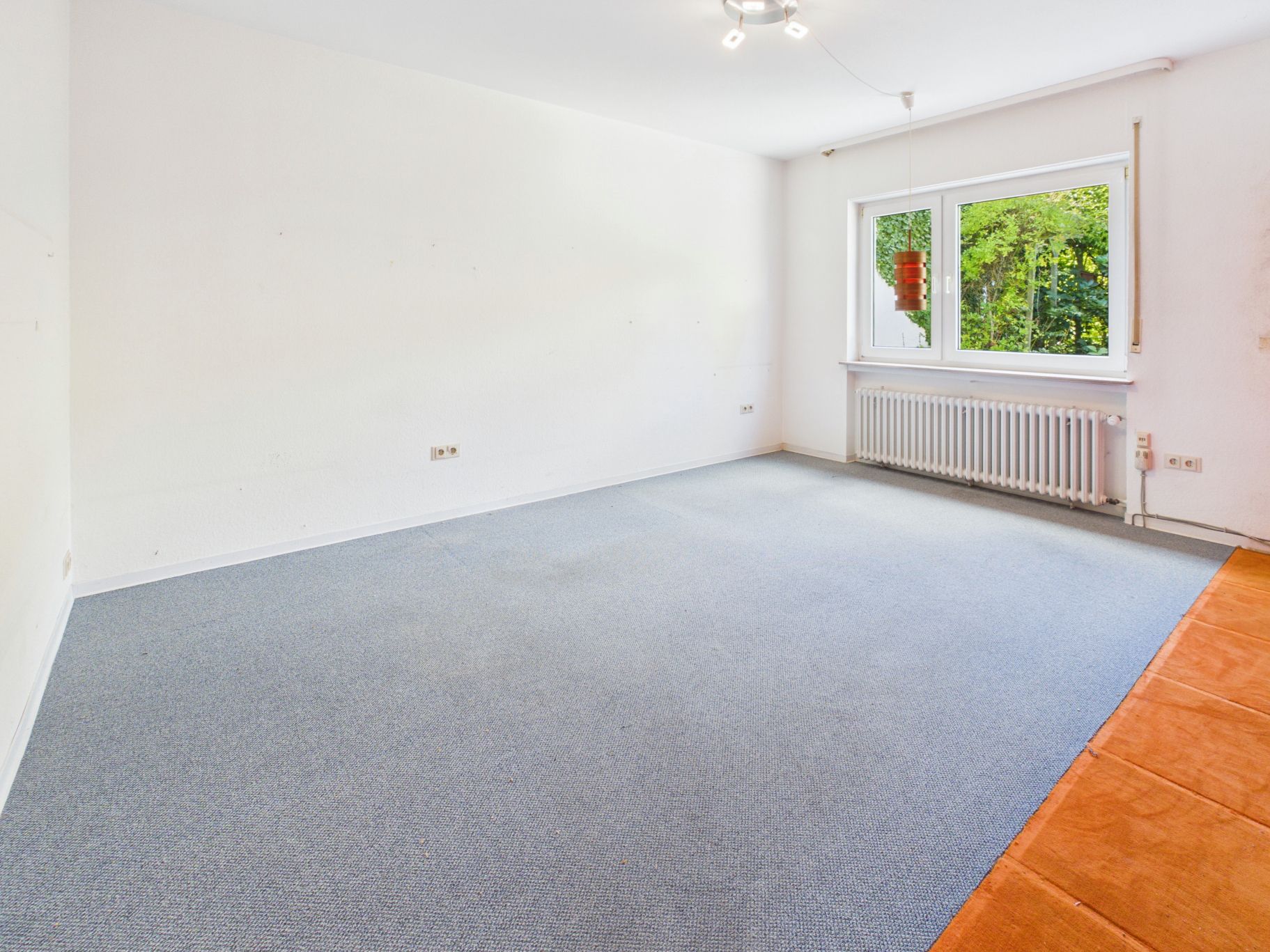 Leerer Raum mit grauem Teppich, großem Fenster und Heizkörper unter dem Fenster.