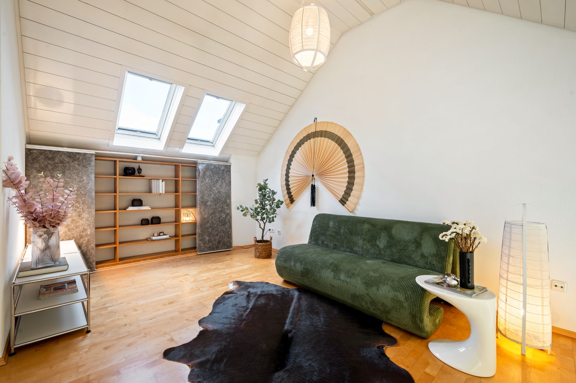 Wohnzimmer mit grünem Sofa, großem Fächer an der Wand, Teppich und Regalen unter Dachfenstern.