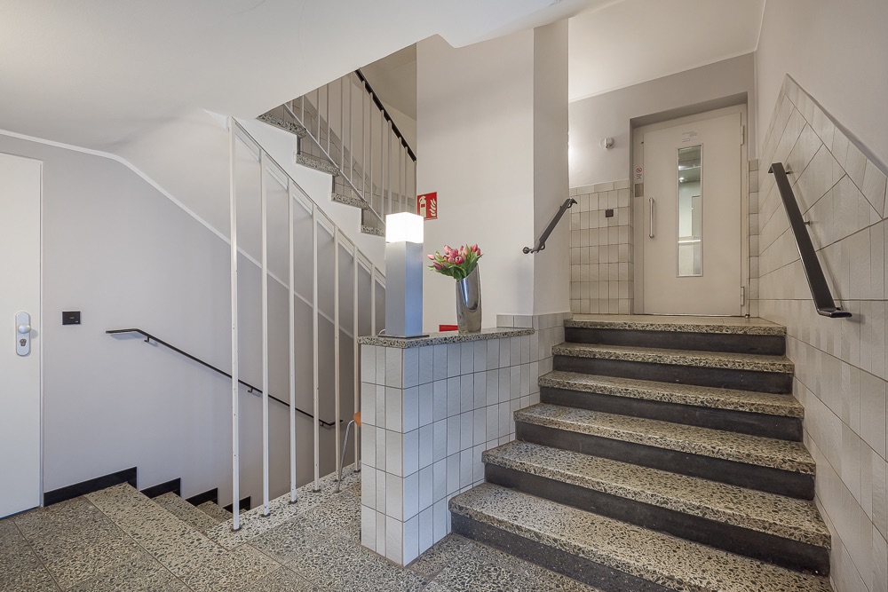 Helles Treppenhaus mit Aufzug, Blumen auf dem Geländer und gefliestem Boden.
