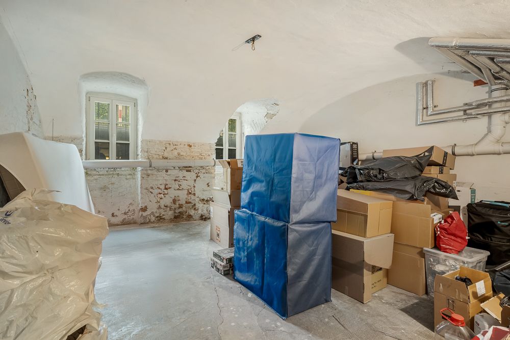 Kellerraum mit sichtbaren Rohren, zwei Fenstern und vielen gestapelten Umzugskartons.