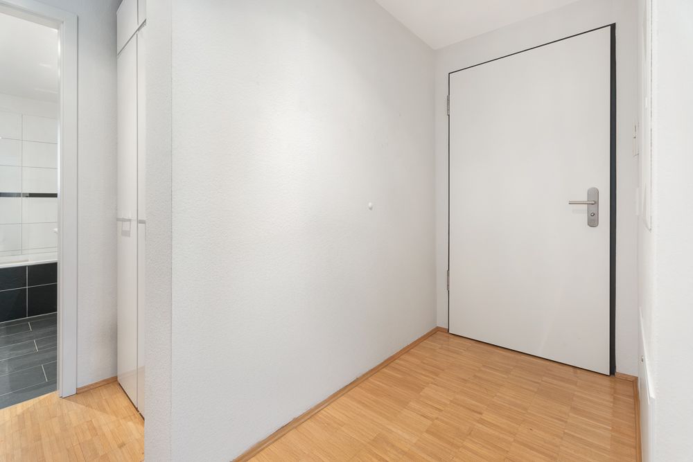 Flur mit Holzboden, weißer Wand, Tür und Zugang zu einem Badezimmer.