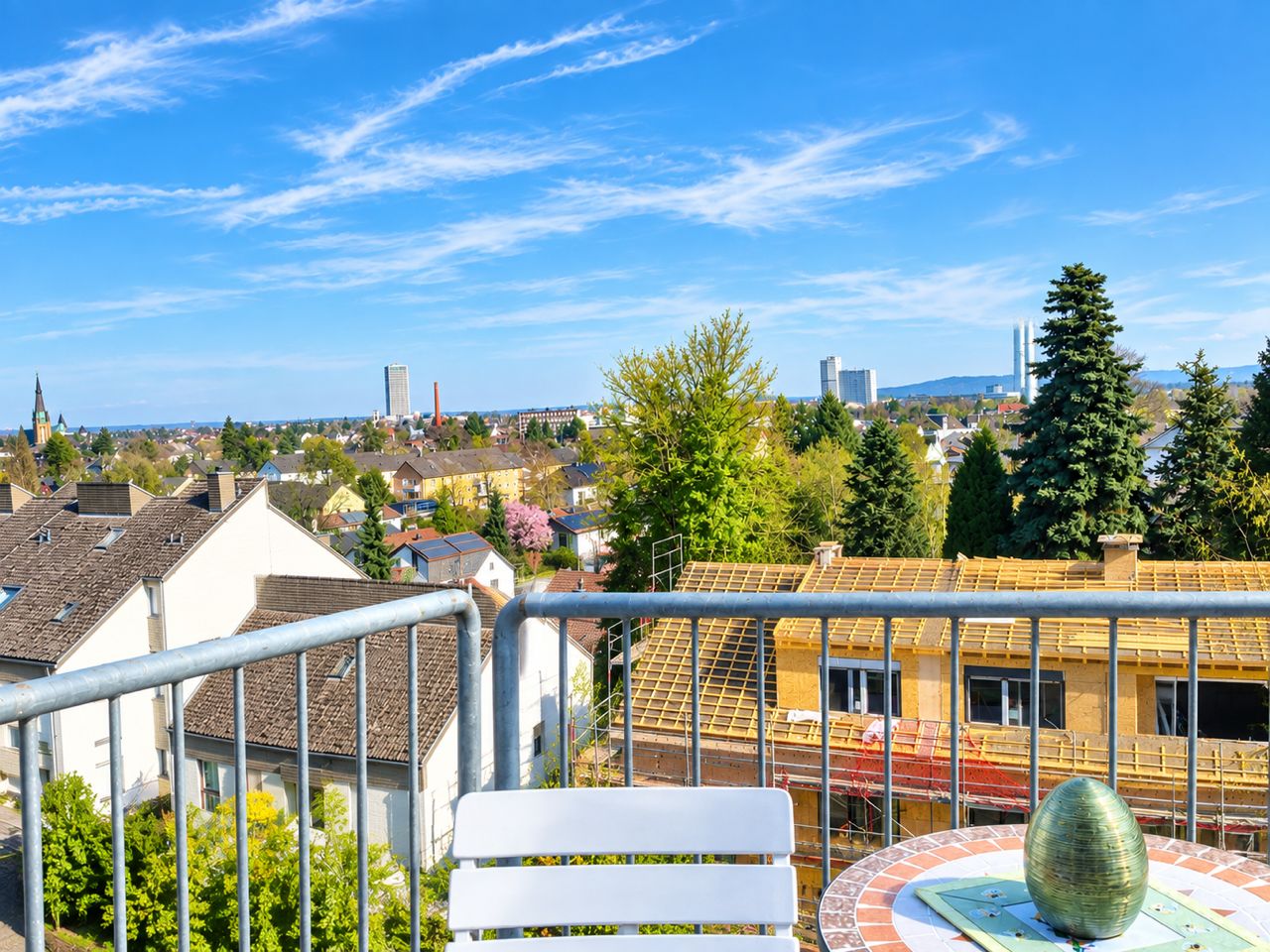 Blick von einem Balkon mit Tisch und Stuhl auf Wohnhäuser, Bäume und blauen Himmel.