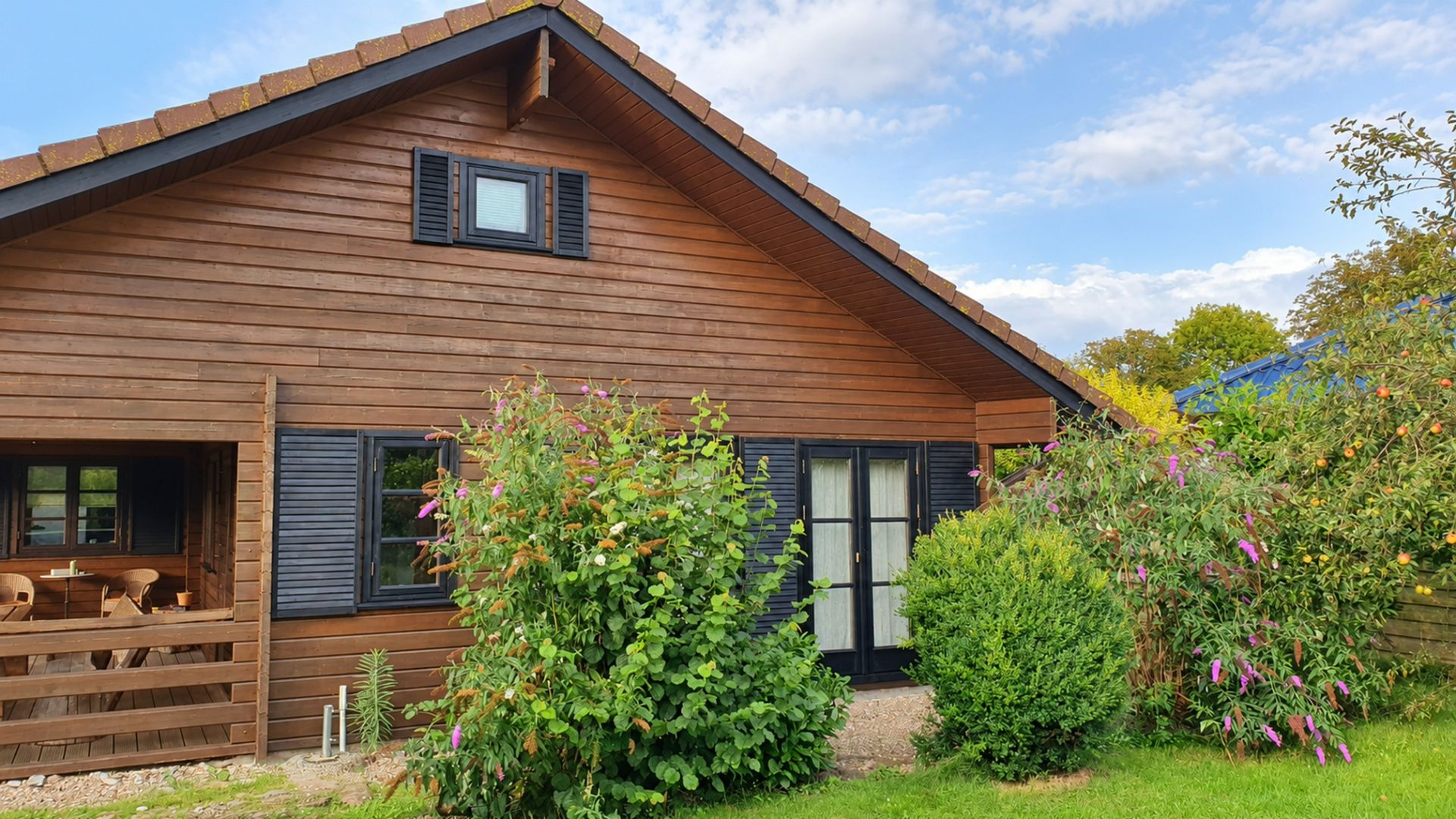Holzhaus mit schwarzen Fensterläden und Garten mit Sträuchern und Büschen im Vordergrund.