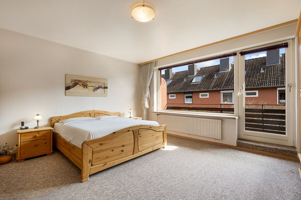 Helles Schlafzimmer mit Holzbett, Nachttischen und großem Fenster mit Blick auf Nachbarhäuser.