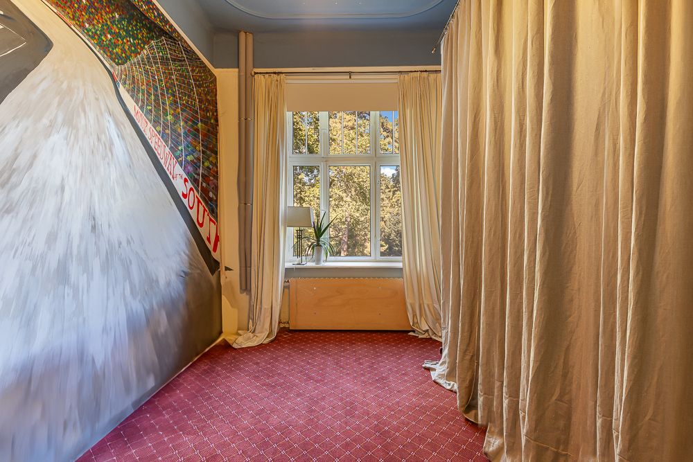 Heller Raum mit rotem Teppich, bemalter Wand, großem Fenster und beigen Vorhängen.