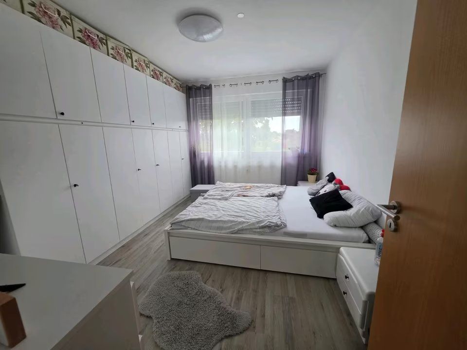 Helles Schlafzimmer mit großem weißen Kleiderschrank, Doppelbett und Fenster mit grauen Vorhängen.