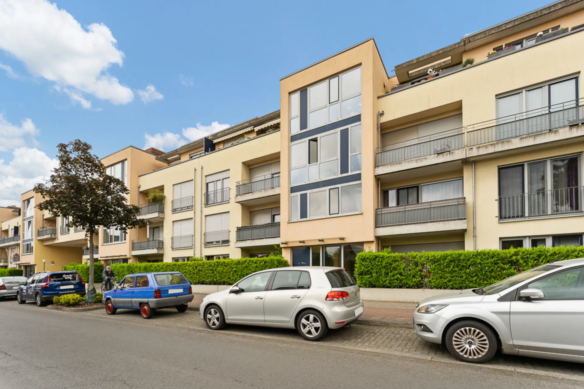 Mehrfamilienhaus mit Balkonen und geparkten Autos an einer ruhigen Straße.