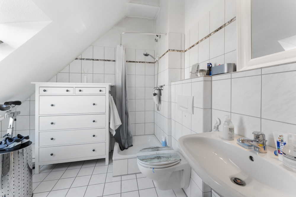 Helles Badezimmer mit Dusche, WC, Waschbecken und weißer Kommode unter einer Dachschräge.