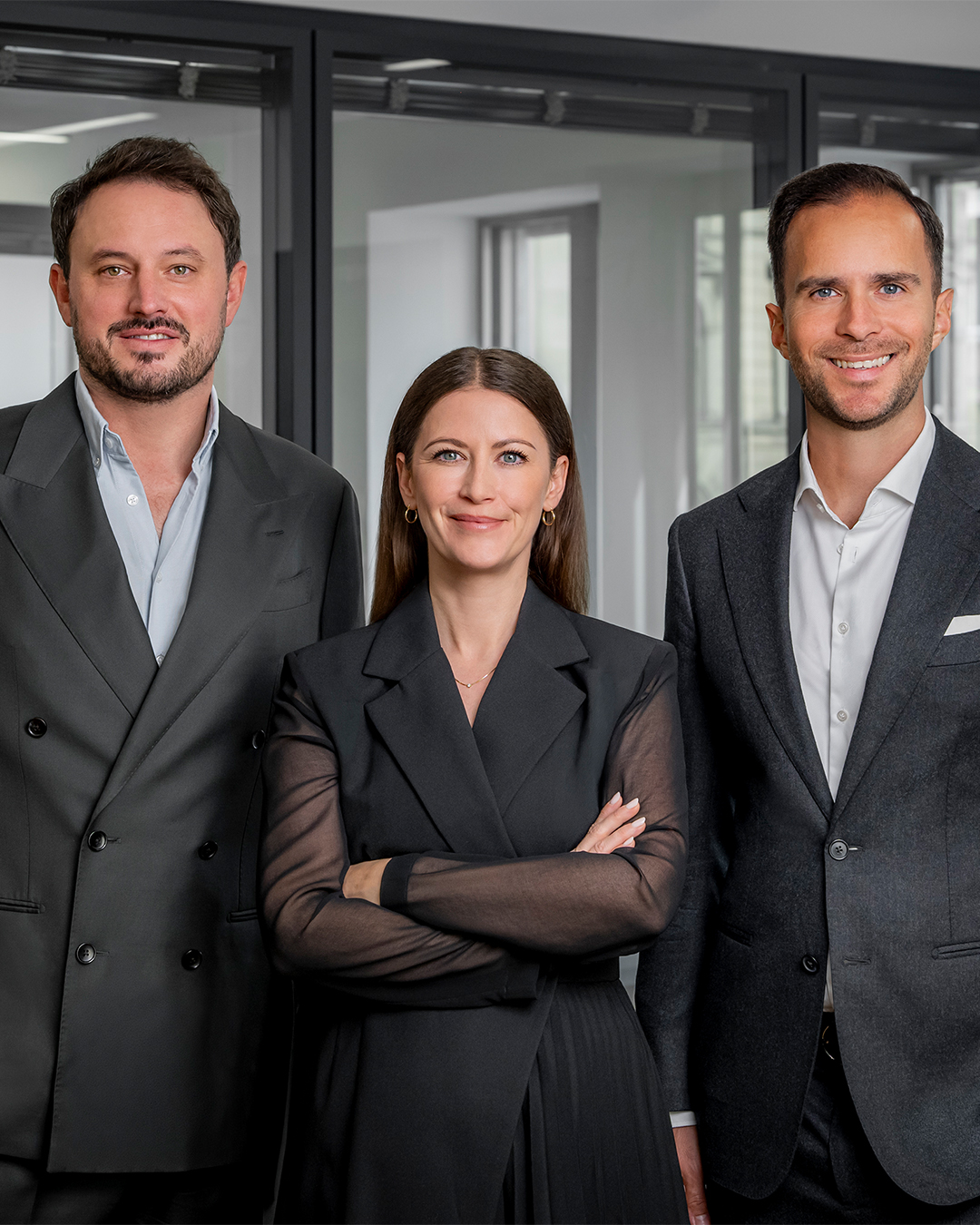 Drei Personen in formeller Kleidung stehen in einem modernen Raum mit Glaswänden.