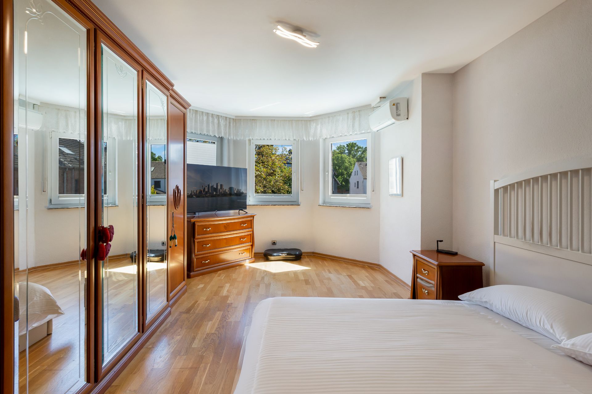 Helles Schlafzimmer mit Holzfußboden, Bett, Kleiderschrank, Kommode und mehreren Fenstern.