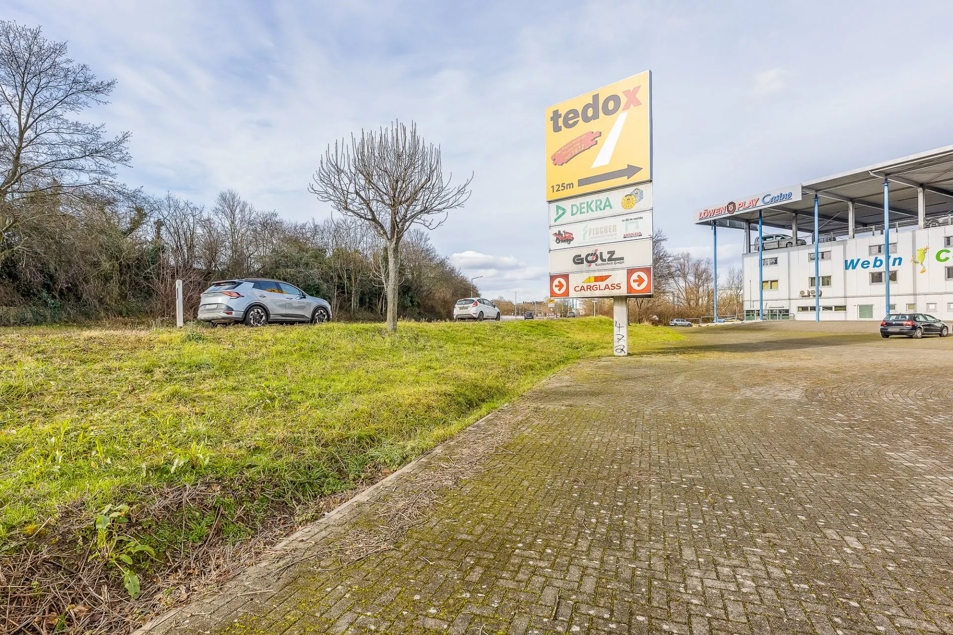 Gepflasterter Parkplatz mit Autos und einem Wegweiser, umgeben von Bäumen und Wiese.