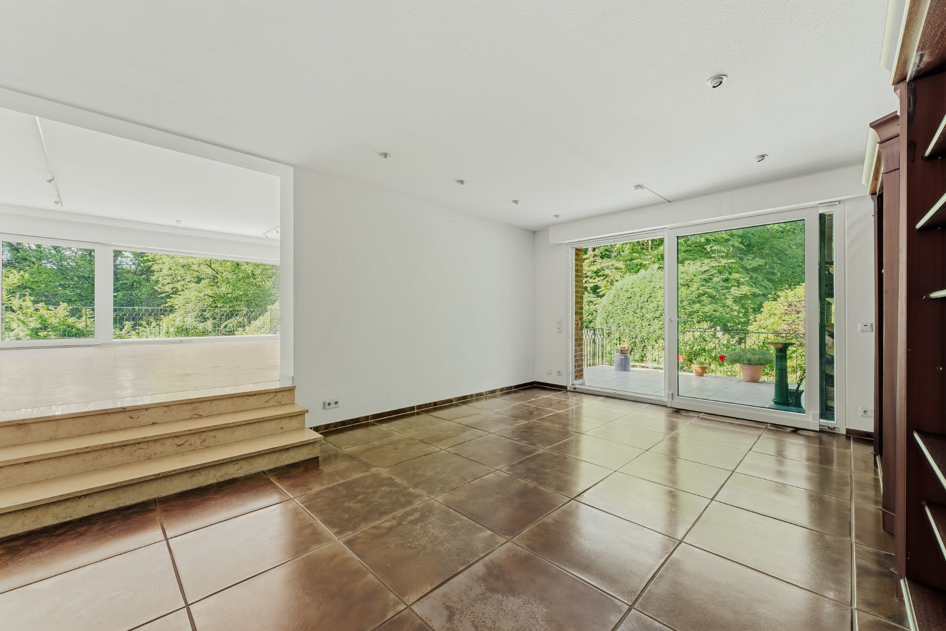 Helles Wohnzimmer mit großen Fenstern und Blick auf eine Terrasse mit Garten, braune Fliesen und Stufen.