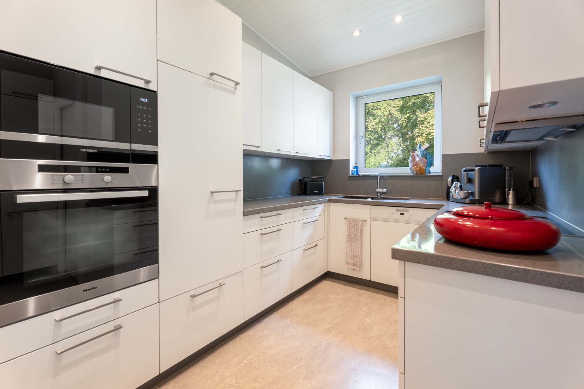 Moderne Küche mit weißen Schränken, großem Fenster und roten Töpfen auf der Arbeitsplatte.