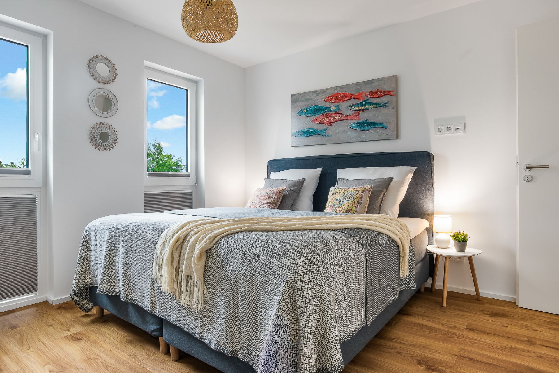 Helles Schlafzimmer mit Doppelbett, zwei Fenstern, Nachttisch und Fischbild an der Wand.