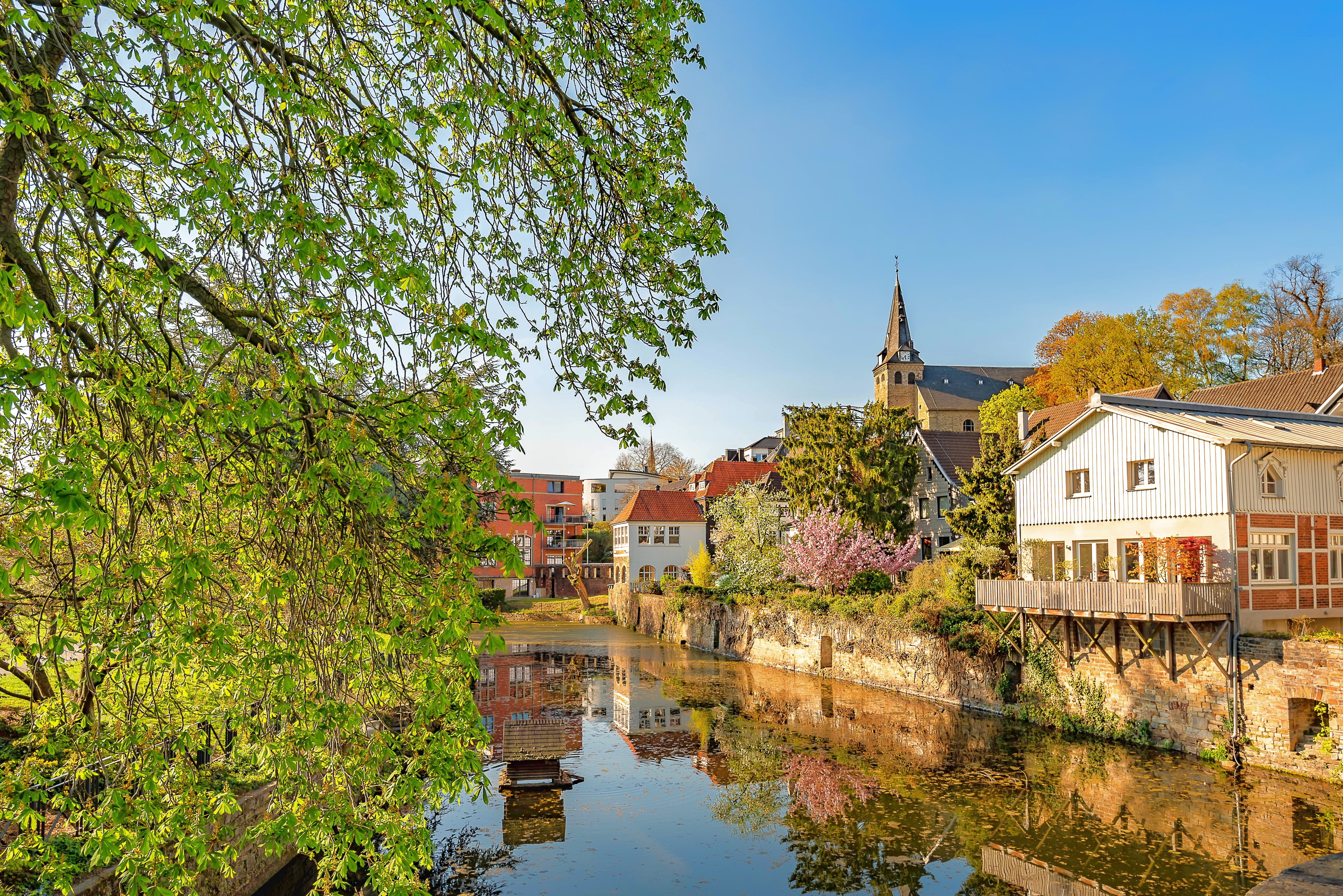 Wohnhäuser und Kirche am Flussufer mit Bäumen und Spiegelung im Wasser.