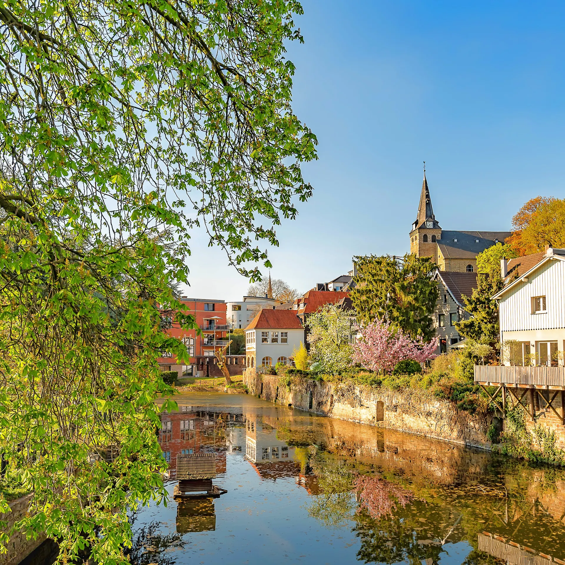 Wohnhäuser und Kirche am Flussufer mit Bäumen und Spiegelung im Wasser.