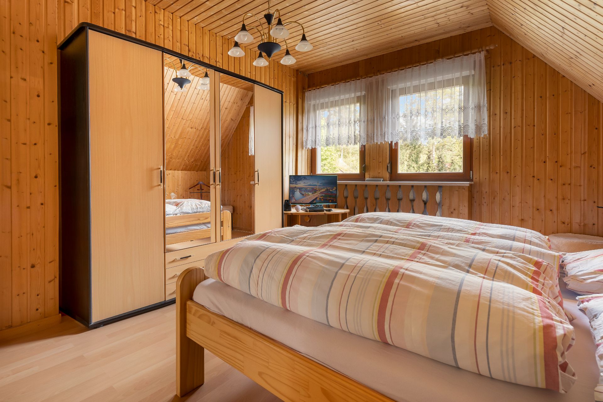 Holzverkleidetes Schlafzimmer mit Doppelbett, Kleiderschrank, Fernseher und zwei Fenstern mit Gardinen.