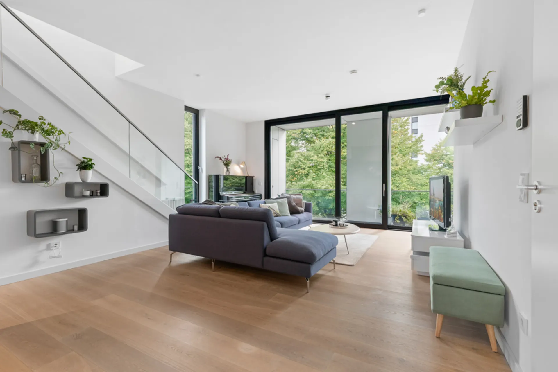 Modernes Wohnzimmer mit Ecksofa, Fernseher, großen Fenstern und Zugang zum Balkon.