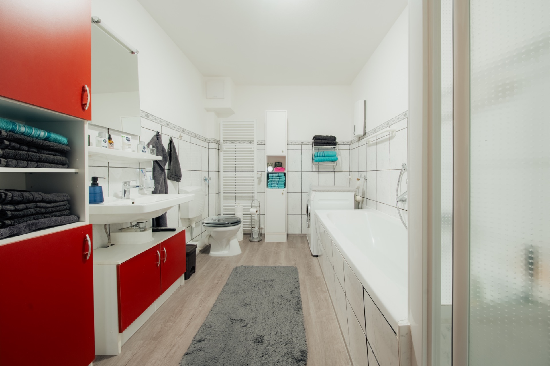 Modernes Bad mit rotem Schrank, Badewanne, Waschbecken, Toilette und grauem Teppich.