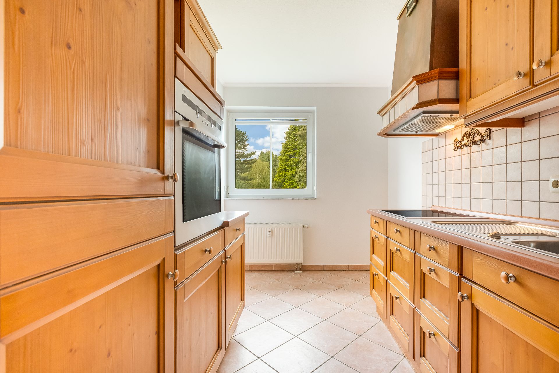 Helle Küche mit Holzfronten, Einbauherd, Fliesenboden und Fenster mit Blick ins Grüne.