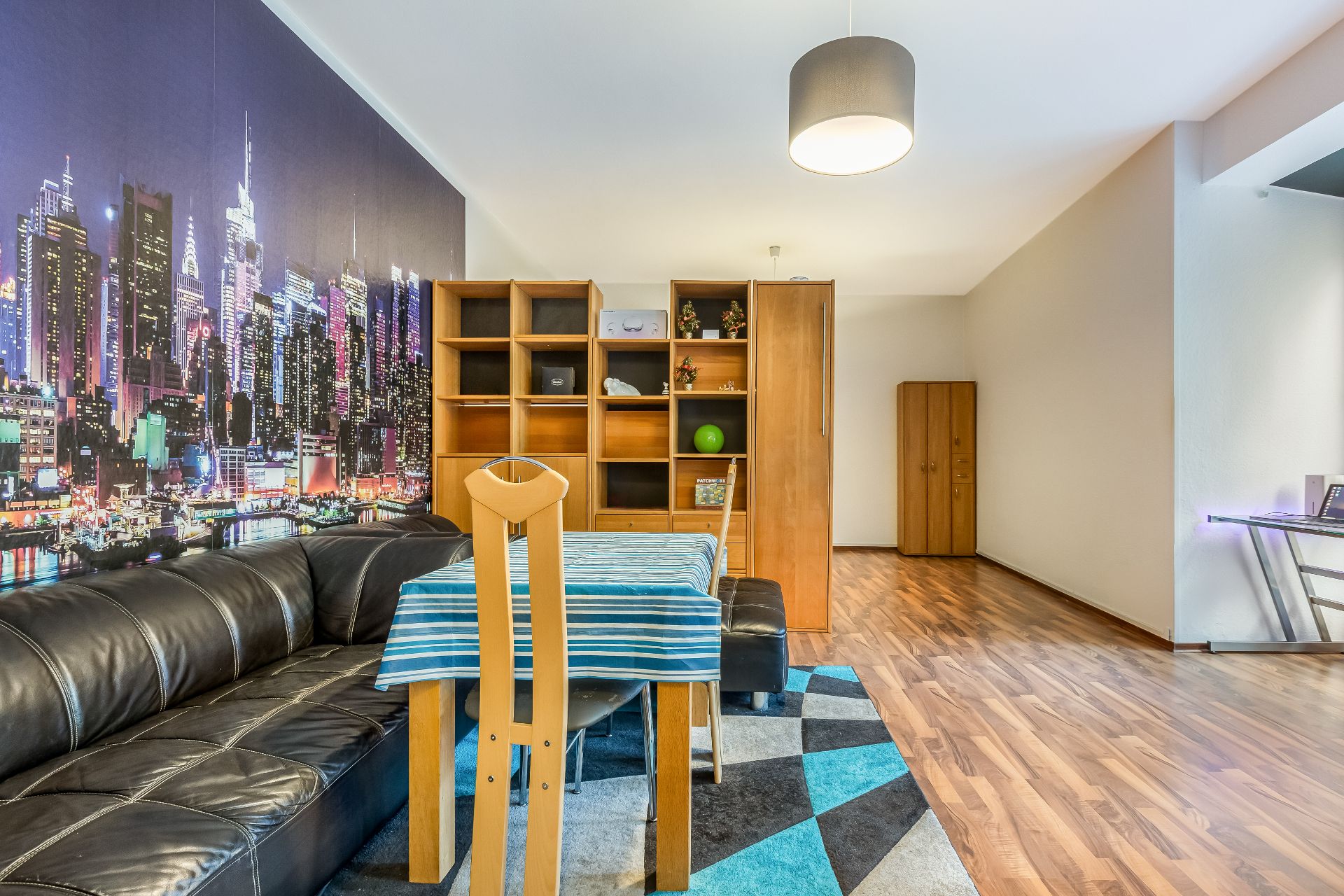 Wohnzimmer mit schwarzem Sofa, Esstisch, Holzmöbeln und Stadtbild-Wandbild.