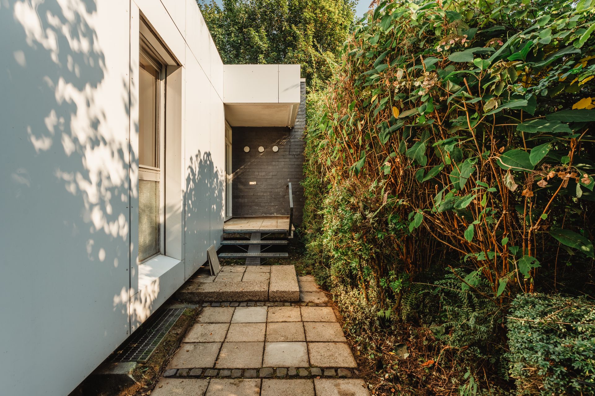 Gepflasterter Weg führt zu einer Tür an einem modernen Wohnhaus, umgeben von dichter Hecke.