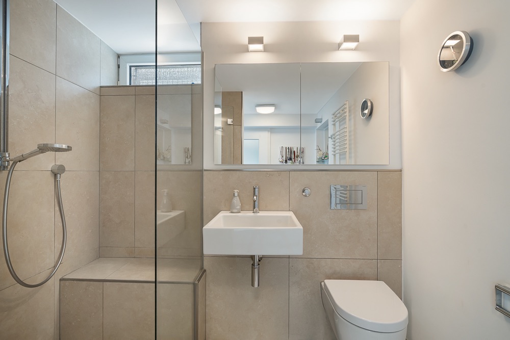 Modernes Badezimmer mit Dusche, Waschbecken und WC, hell gefliest und mit Spiegel über dem Waschbecken.