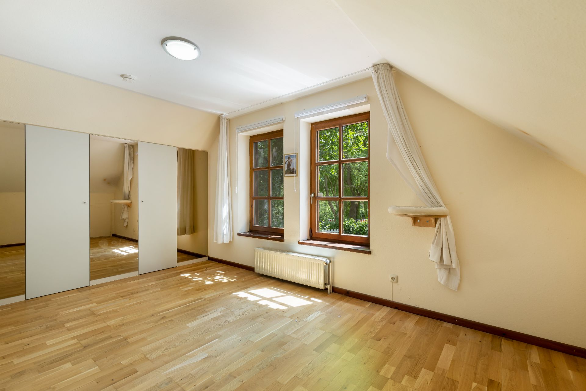 Helles Zimmer mit zwei Fenstern, Holzfußboden und Einbauschrank mit Spiegeltüren.