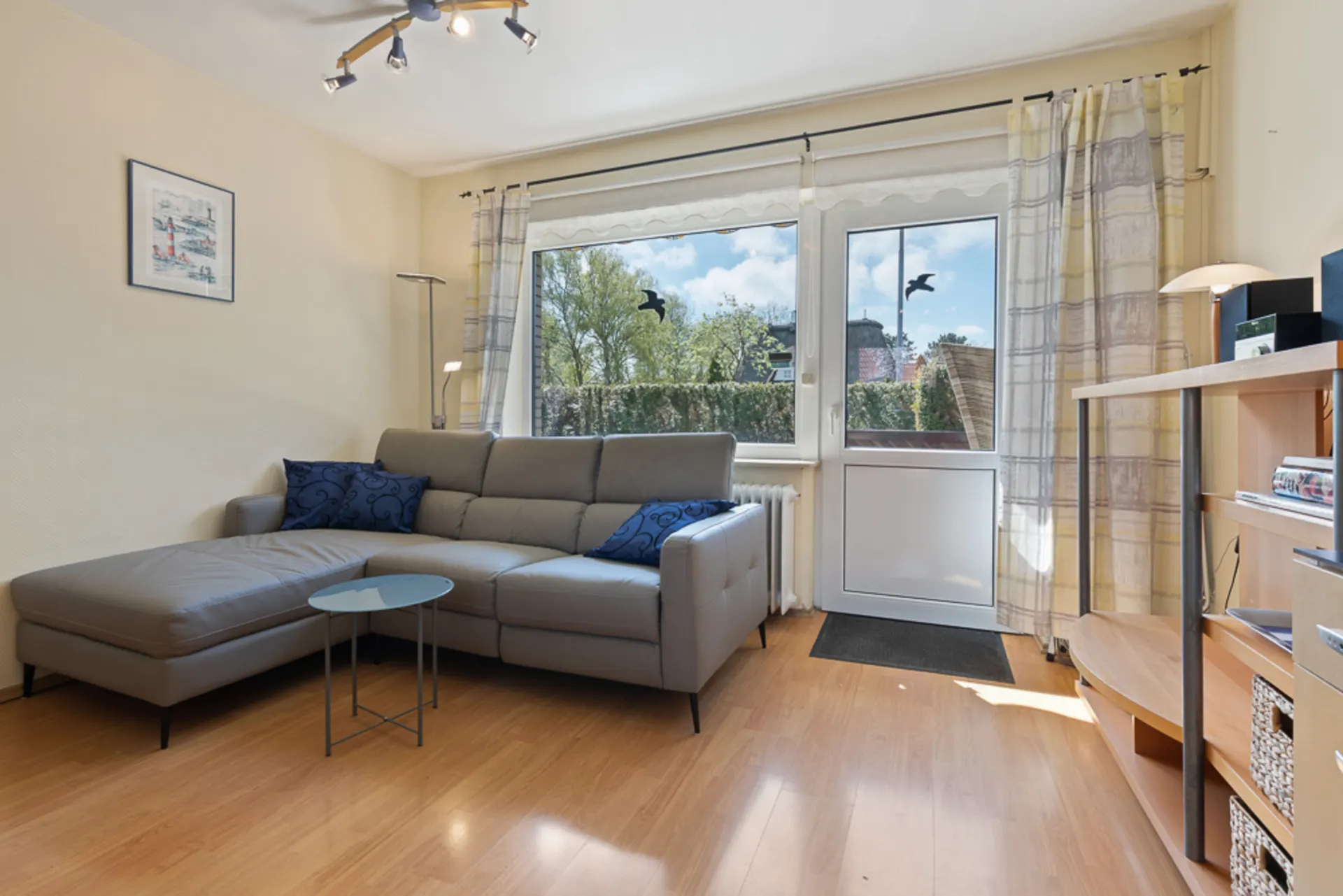 Helles Wohnzimmer mit grauem Ecksofa, Couchtisch, Regal und Zugang zum Balkon.