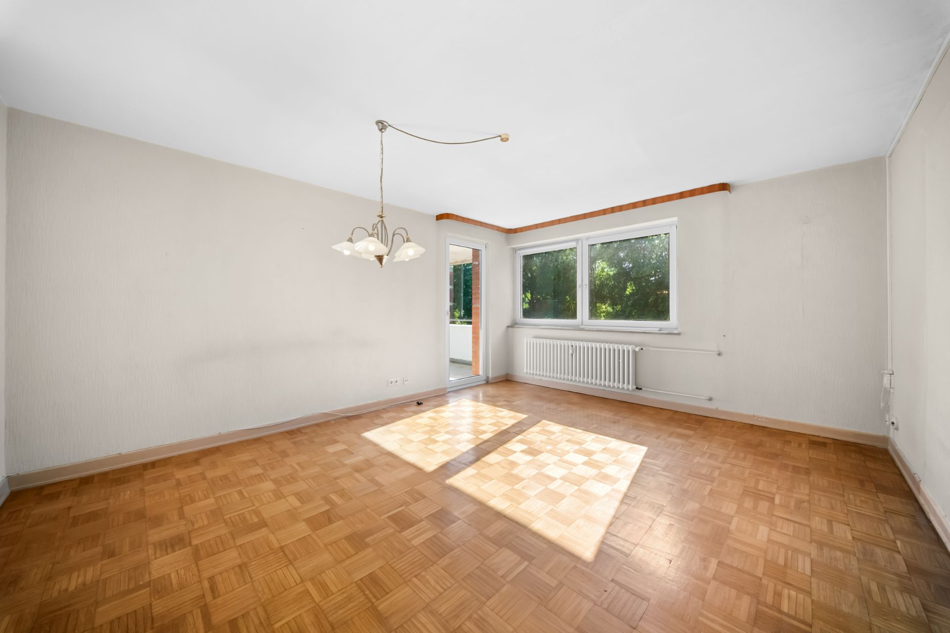 Heller leerer Raum mit Parkettboden, zwei Fenstern, Heizkörper und Deckenlampe.
