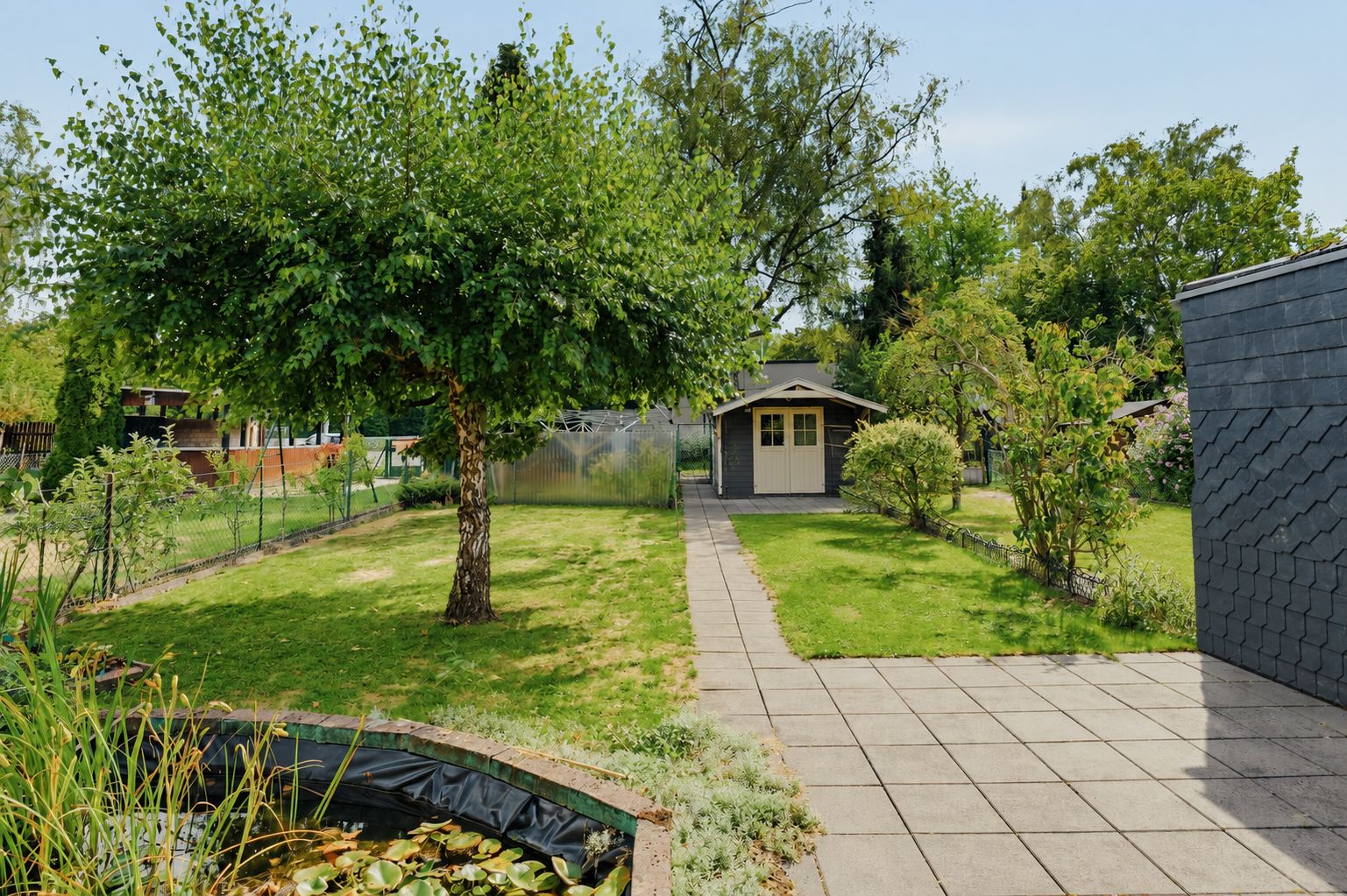 Gepflegter Garten mit Teich, Baum, Weg und kleinem Gartenhaus im Hintergrund.