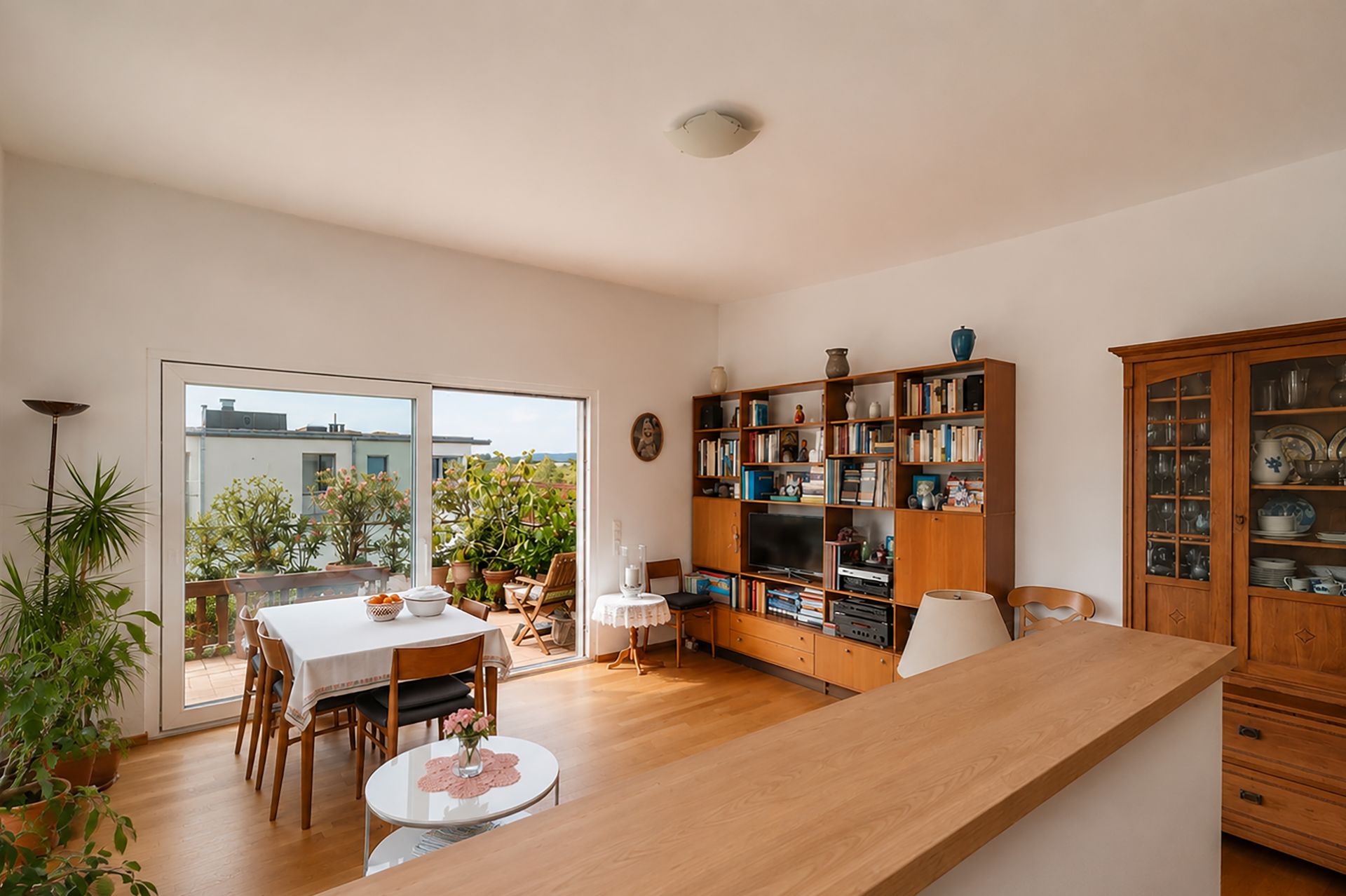Helles Wohnzimmer mit Esstisch, Bücherregal, Vitrine und Zugang zu einem Balkon mit Pflanzen.