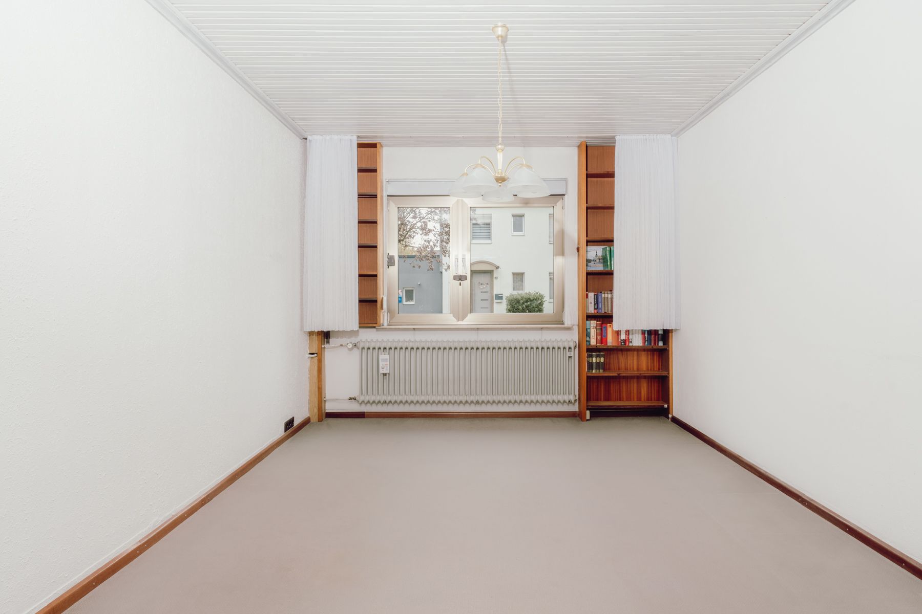 Leeres Zimmer mit Teppichboden, Fenster, zwei Bücherregalen und einem Deckenleuchter.